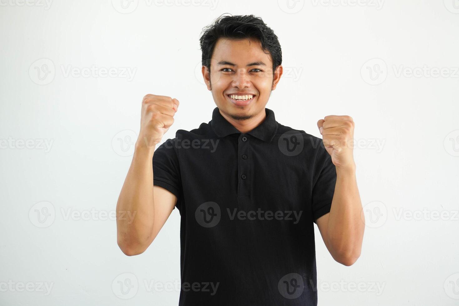 smiling or happy young asian man clenched his fists cheering carefree and excited. Victory concept, wearing black polo t shirt isolated on white background photo