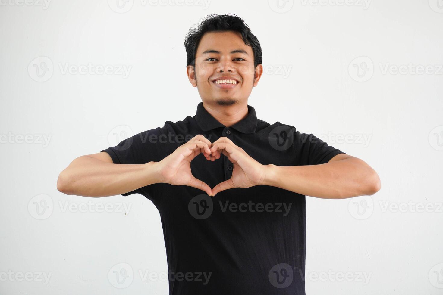 young asian man posing on a white backdrop smiling and showing a heart shape with hands, wearing black polo t shirt. photo