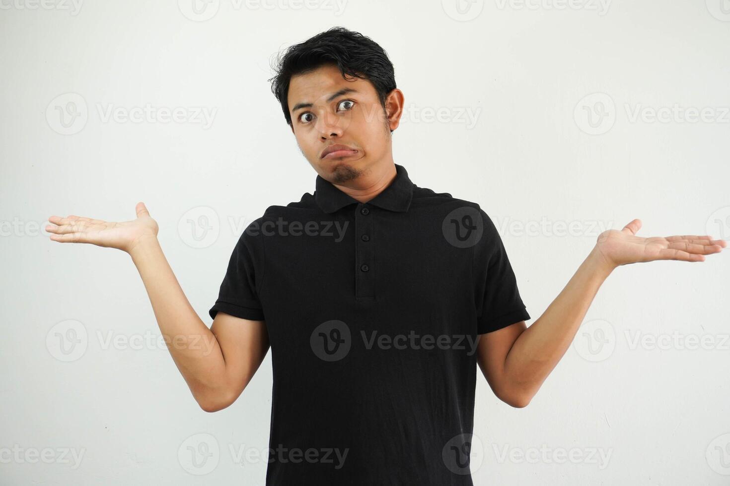 young asian man posing on a white backdrop confused and doubtful shrugging shoulders to hold a copy space, wearing black polo t shirt. photo