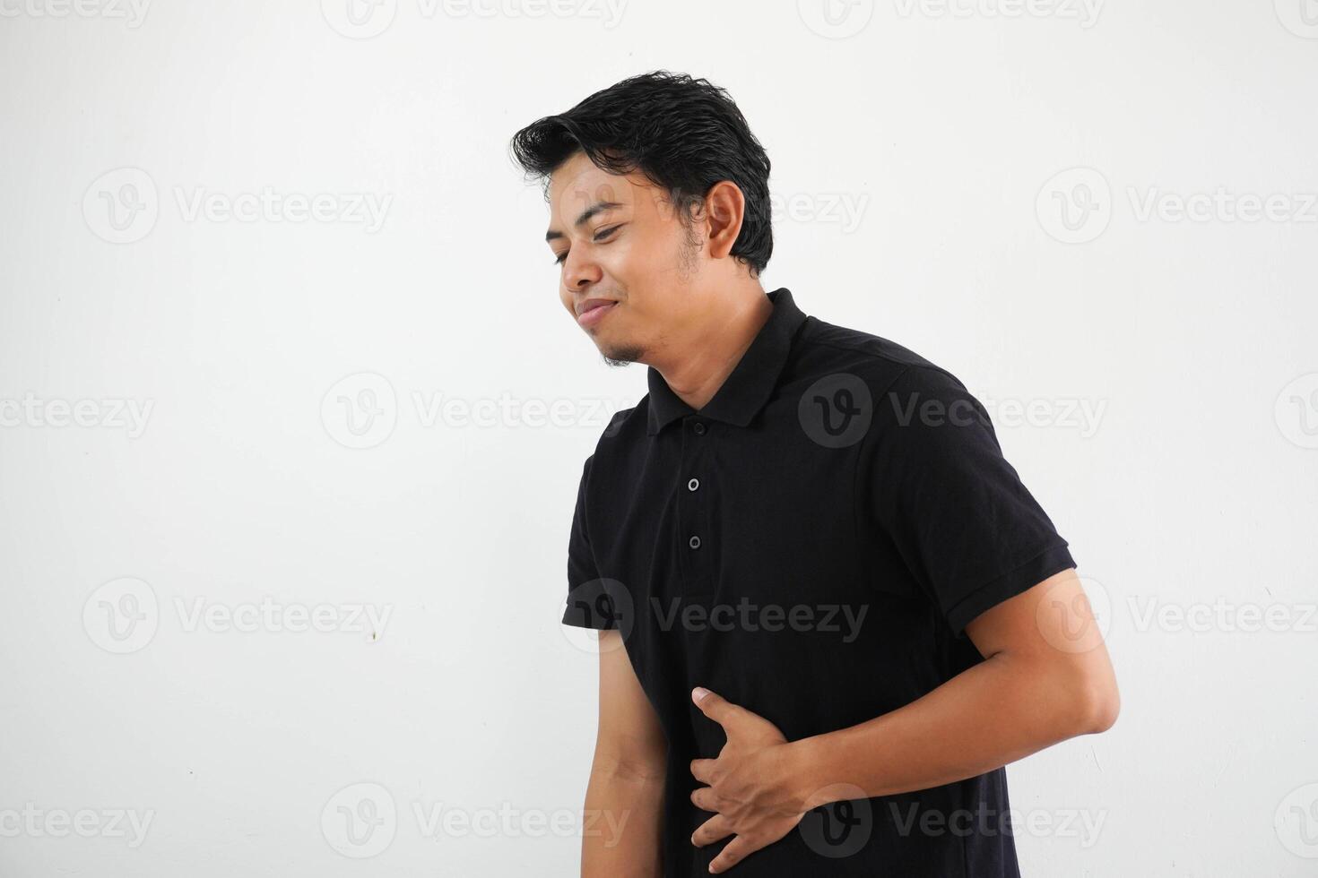 young asian man posing on a white backdrop wearing black polo t shirt touches tummy, smiles gently, eating and satisfaction concept. photo