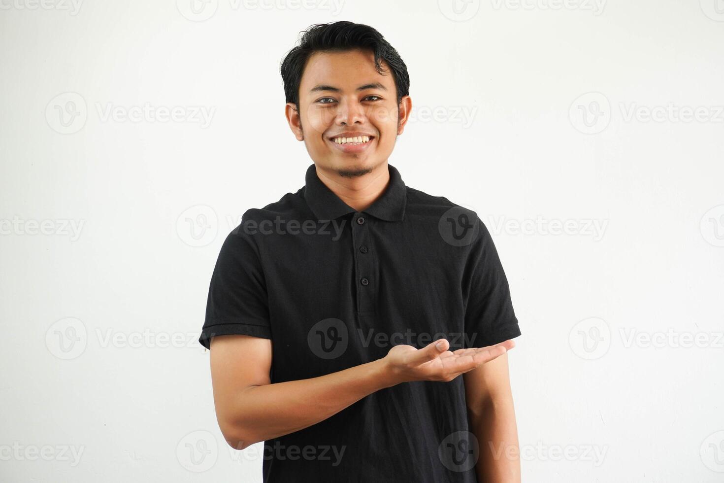 smiling young asian man in black polo shirt, white backdrop showing a copy space on a palm and holding another hand on waist. photo