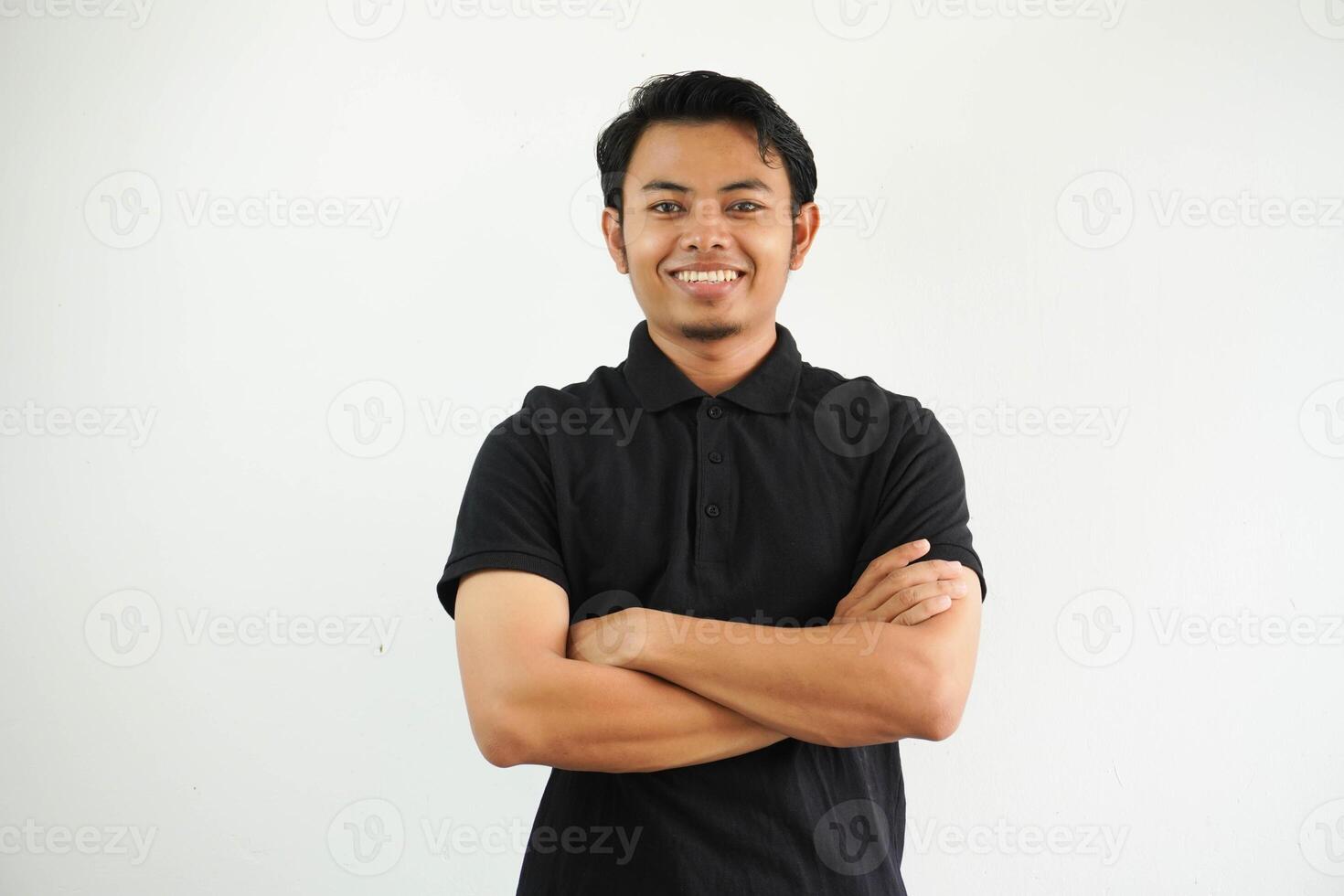 joven asiático hombre sonriente contento a el cámara con brazos cruzado vistiendo negro polo t camisa aislado en blanco antecedentes foto