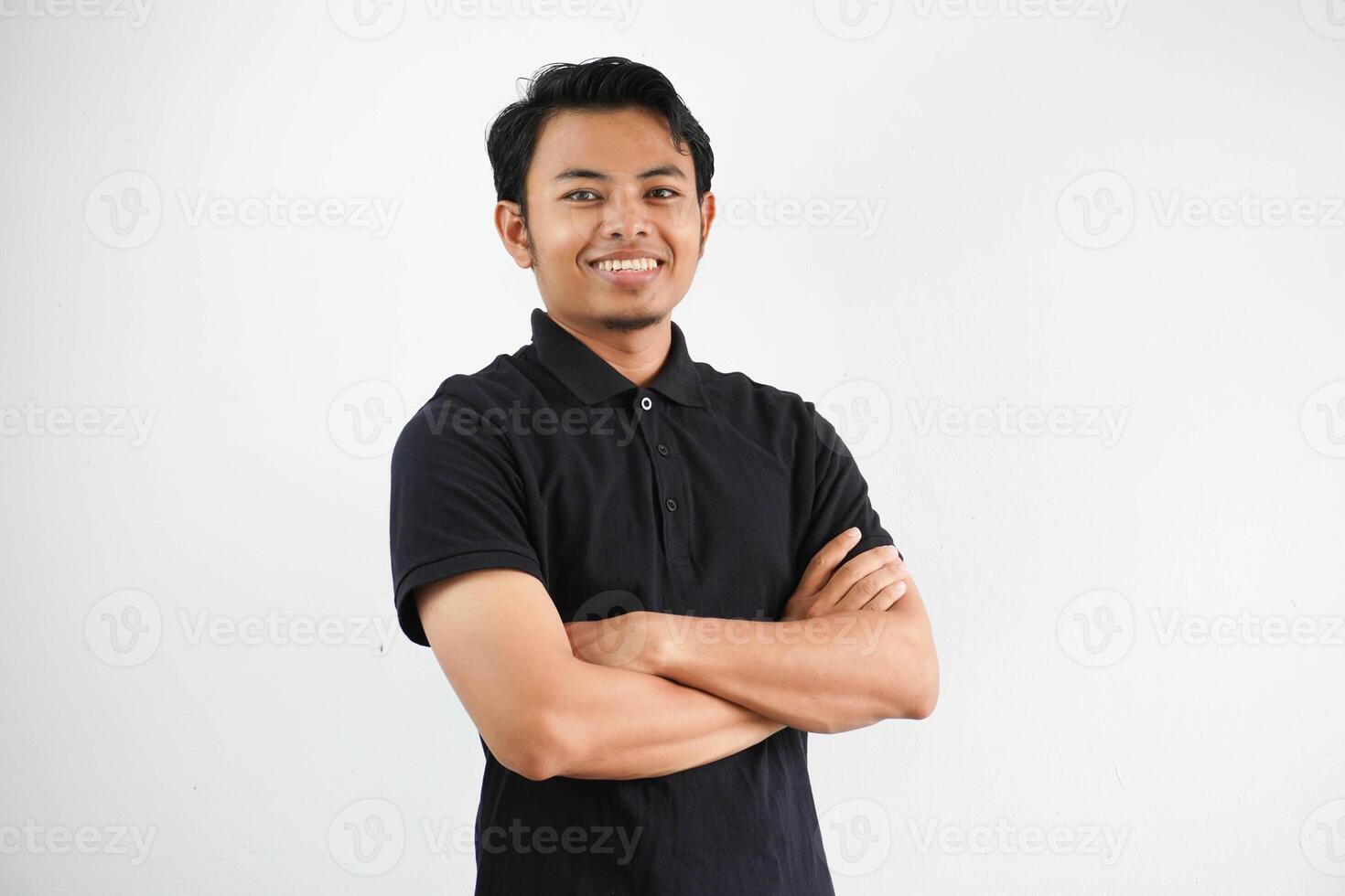 joven asiático hombre sonriente contento a el cámara con brazos cruzado vistiendo negro polo t camisa aislado en blanco antecedentes foto