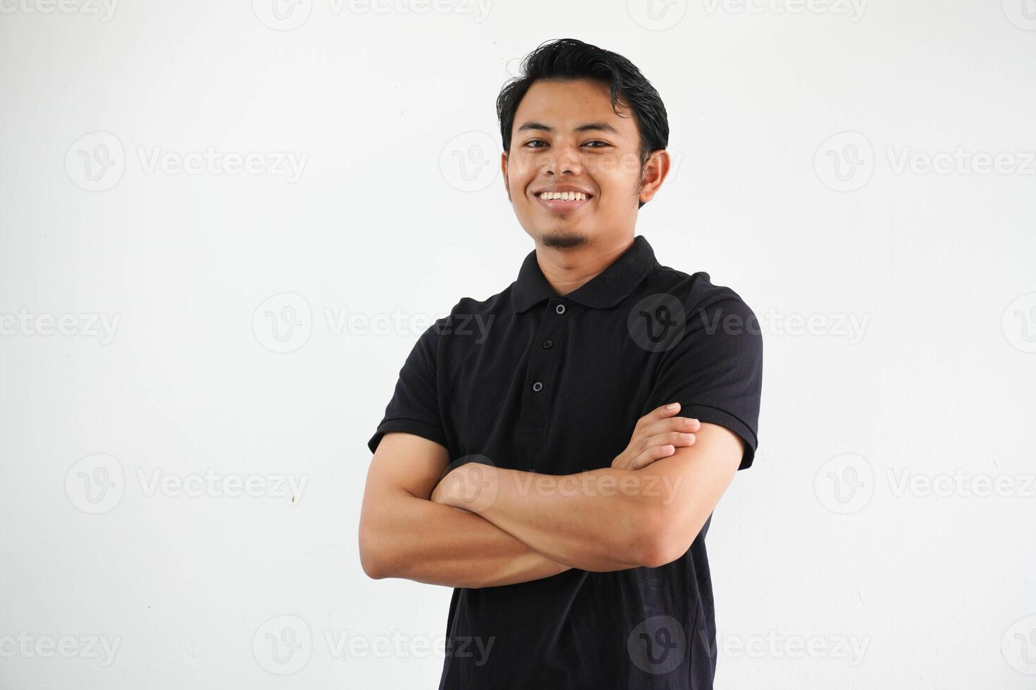 joven asiático hombre sonriente contento a el cámara con brazos cruzado vistiendo negro polo t camisa aislado en blanco antecedentes foto