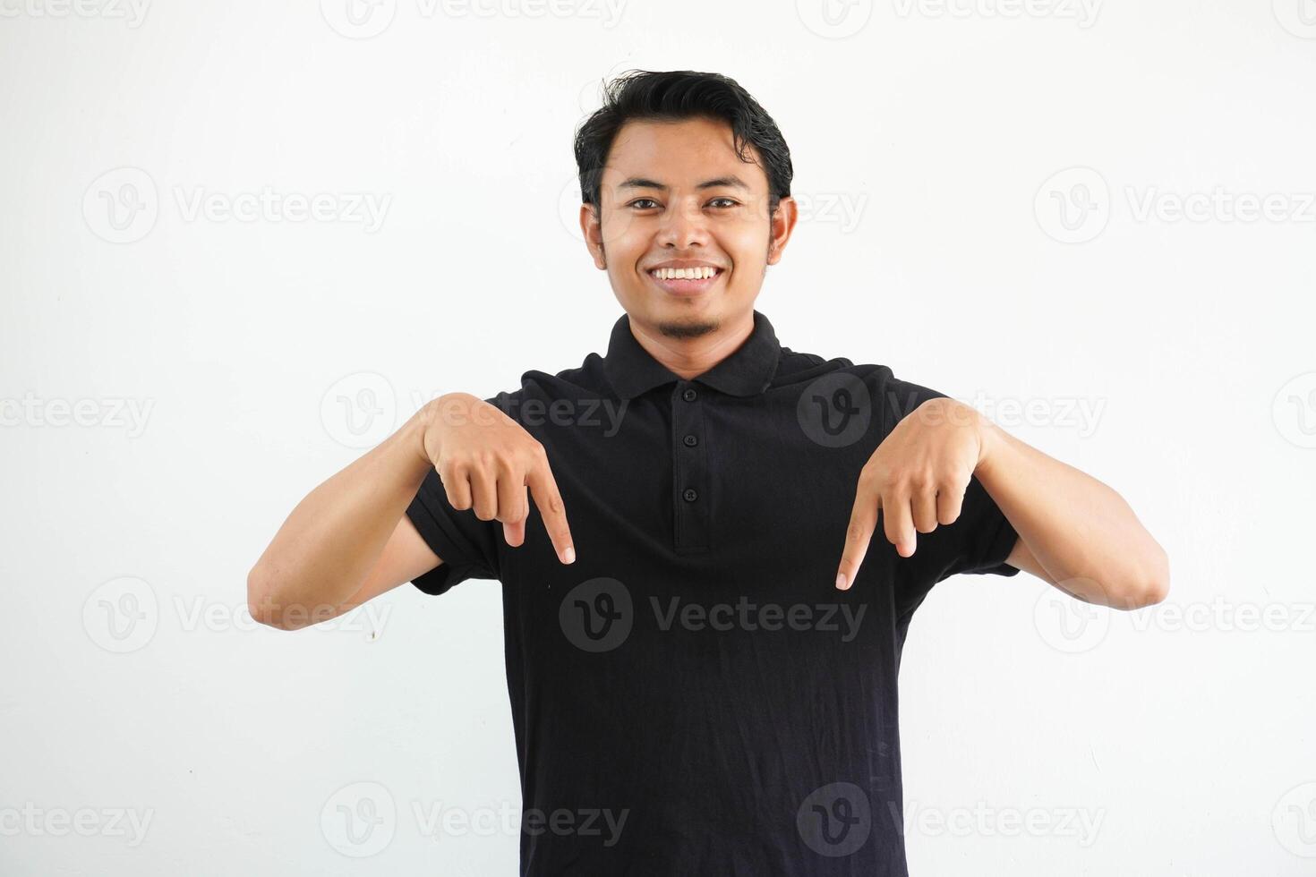 young asian man smiling at the camera and pointing below wearing black polo t shirt isolated on white background. photo