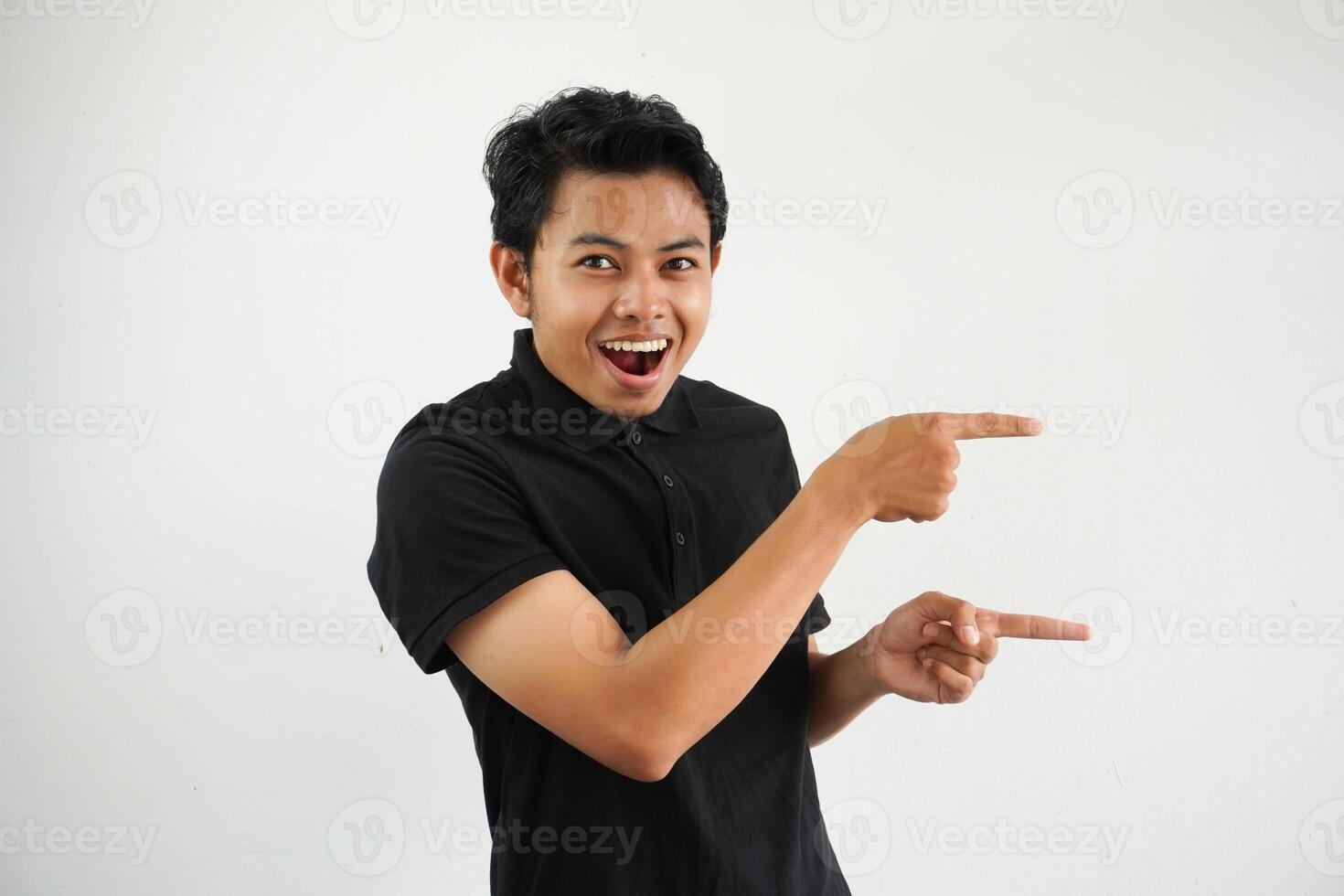 emocionado joven asiático hombre sonriente confidente con ambos mano señalando a el izquierda lado vistiendo negro polo t camisa aislado blanco antecedentes foto