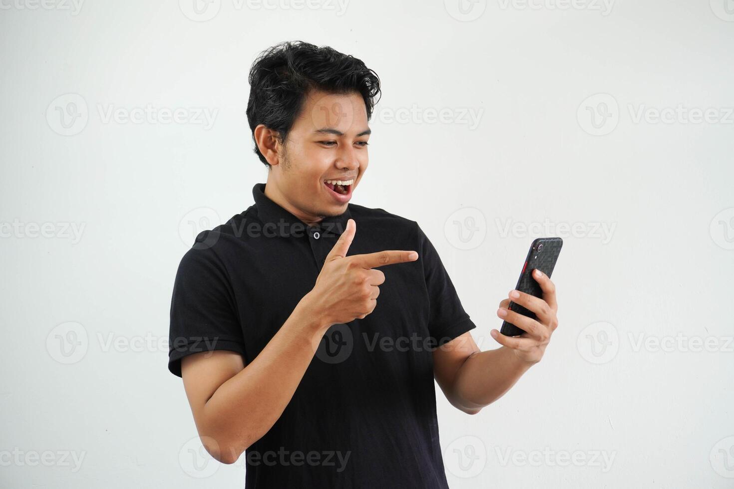excited young asian man smiling happy while pointing to mobile phone that he hold wearing black polo t shirt isolated on white background photo