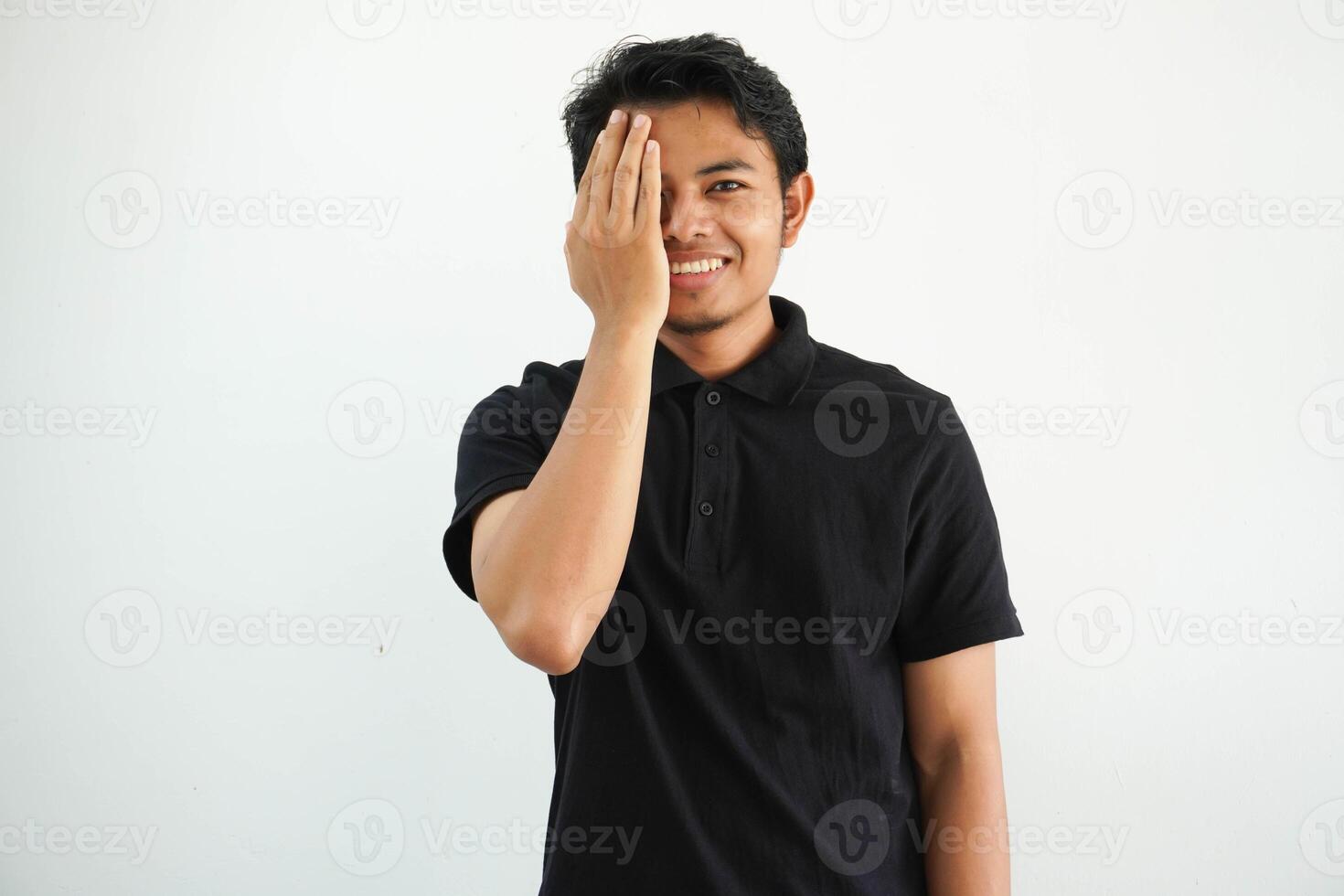 young asian man posing on a white backdrop having fun covering half of face with palm, wearing black polo t shirt. photo