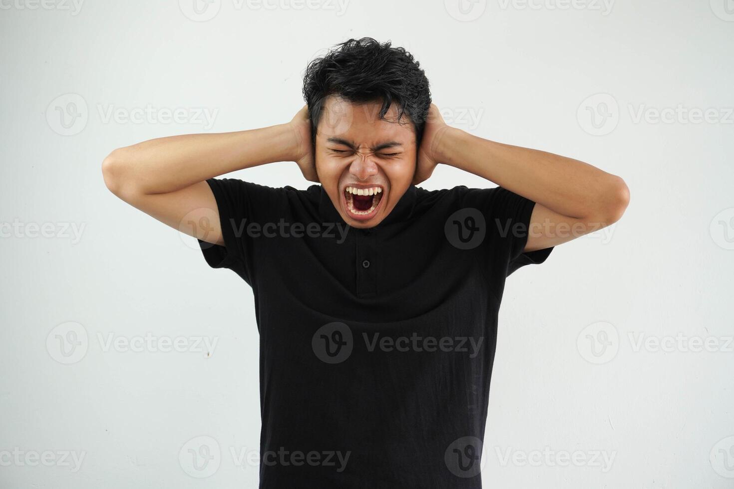 joven asiático hombre gritando con abierto boca y manos cubierta orejas, enojado expresión en contra algo, vistiendo negro polo t camisa aislado en blanco antecedentes foto