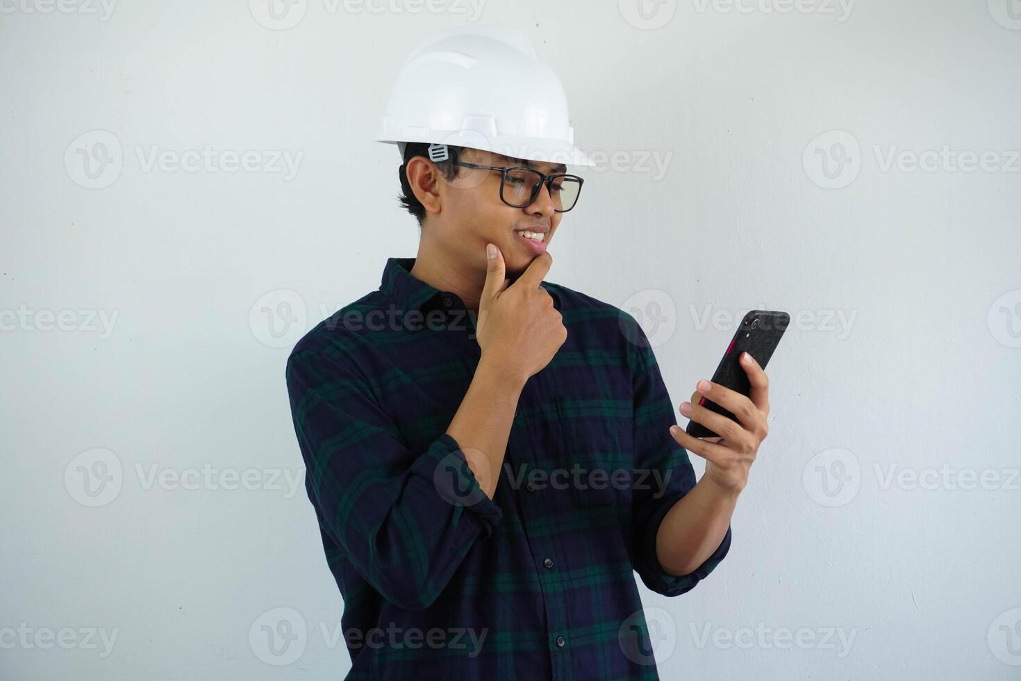 young asian male architect engineer smiling holding his chin thinking while looking at the cellphone he was holding isolated on white background. photo