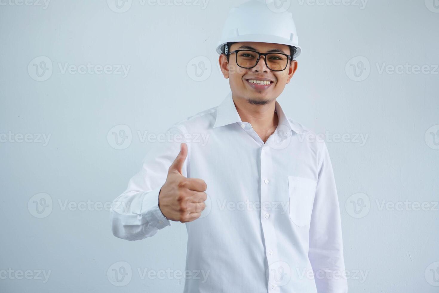 Young asian architect man wearing builder safety helmet over isolated background doing happy thumbs up gesture with hand. Approving expression looking at the camera showing success. photo