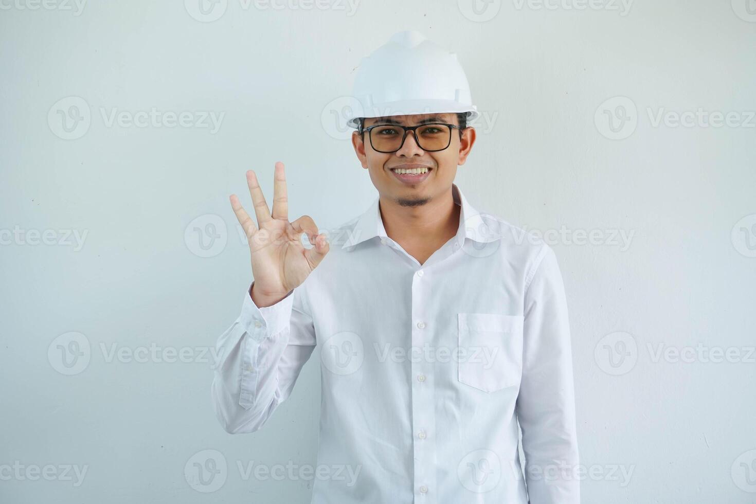 smiling young Asian male engineer wearing white hard hat making ok sign with hand for construction work isolated on white background, copy space. photo