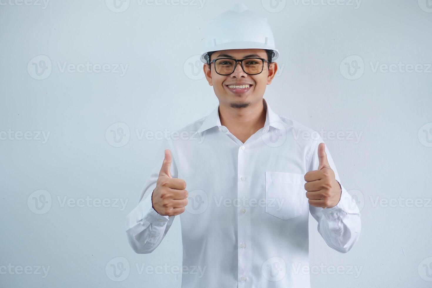 Young asian architect man wearing builder safety helmet over isolated background doing happy thumbs up gesture with hand. Approving expression looking at the camera showing success. photo