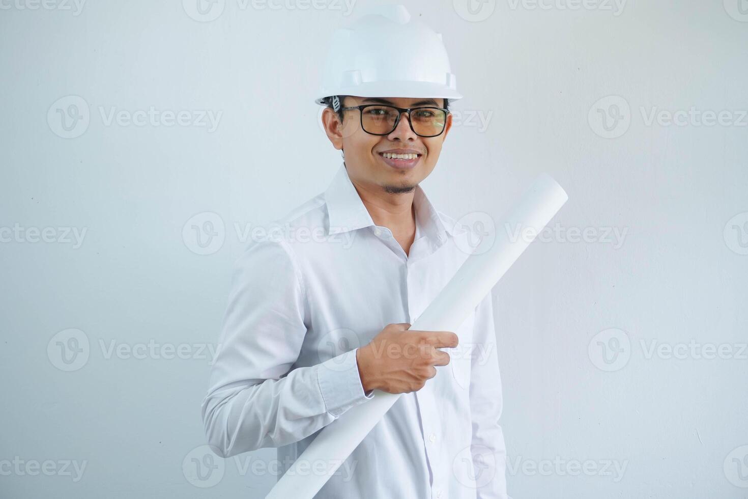 smiling young Asian architect wearing helmet hardhat and holding project paper plan isolated on white background. photo