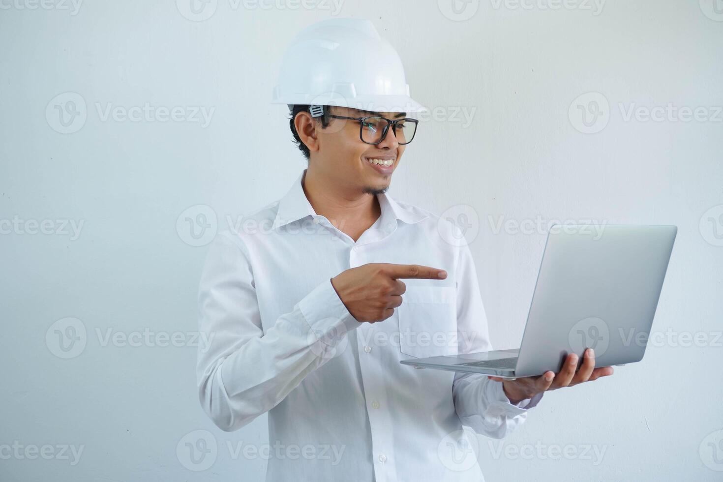 smiling young asian architect man wearing builder safety helmet pointing his laptop isolated on white background photo