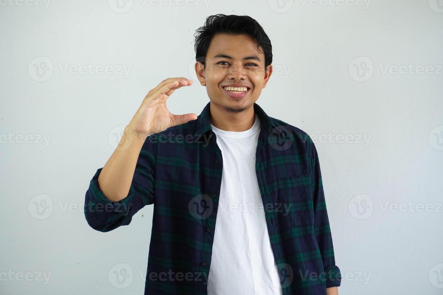 young asian man posing on a white backdrop holding something little with forefingers, smiling and confident. photo