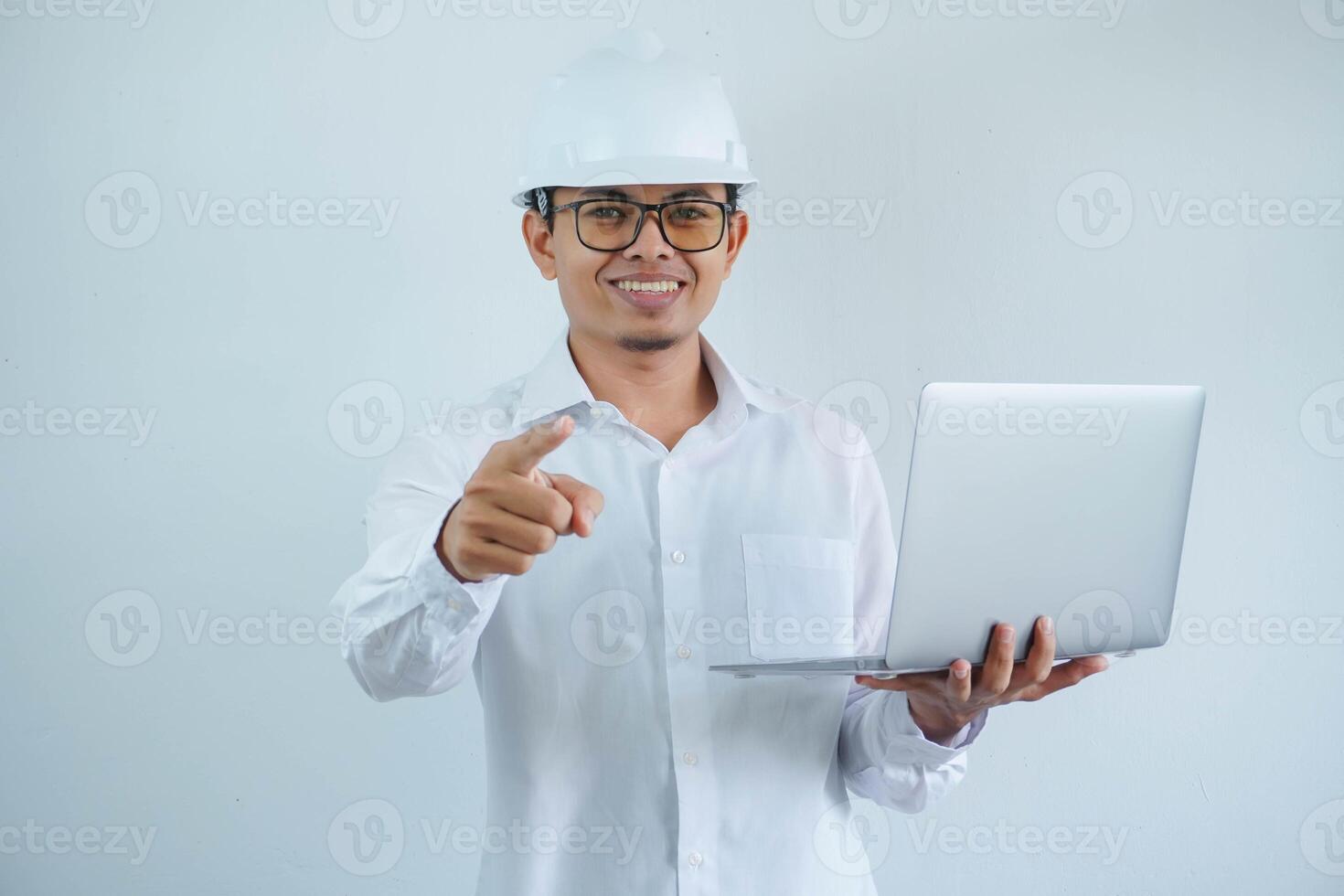 smiling young asian architect man wearing builder safety helmet holding laptop and pointing forward isolated on white background photo