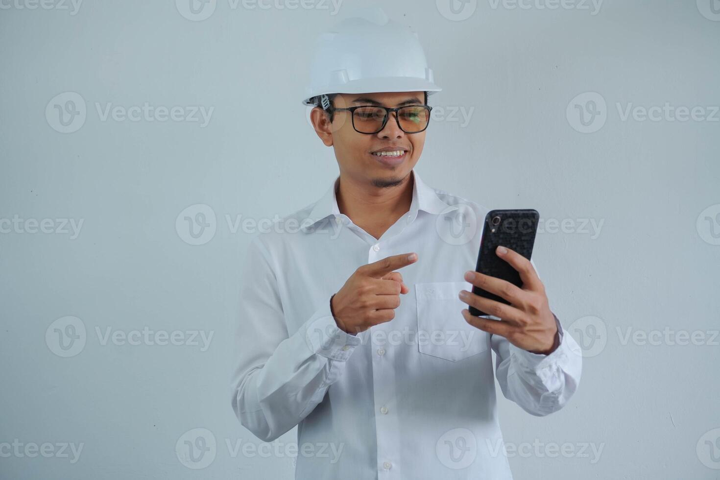 smiling young asian architect man wearing builder safety helmet pointing his mobile phone isolated on white background photo