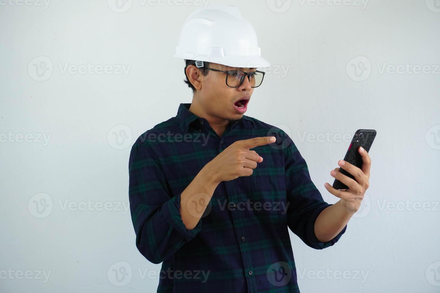 shocked young asian architect man wearing builder safety helmet pointing his mobile phone isolated on white background photo