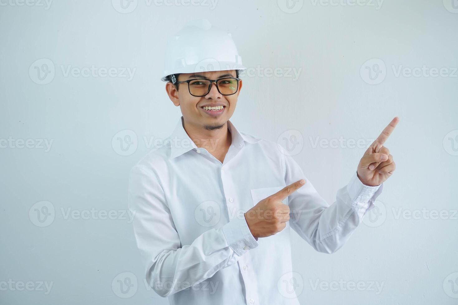 young asian architect man wearing white hard hat safety helmet looking camera with finger pointing the left side isolated on white background photo