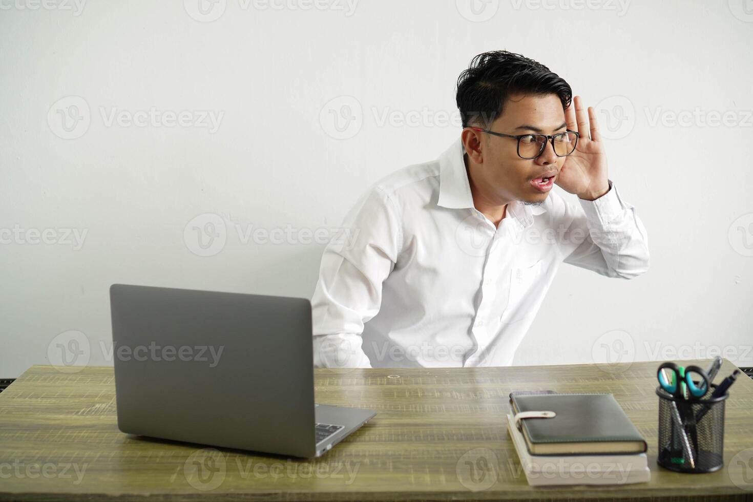 young asian businessman in a workplace listening to something by putting hand on the ear, wear white shirt with glasses isolated photo