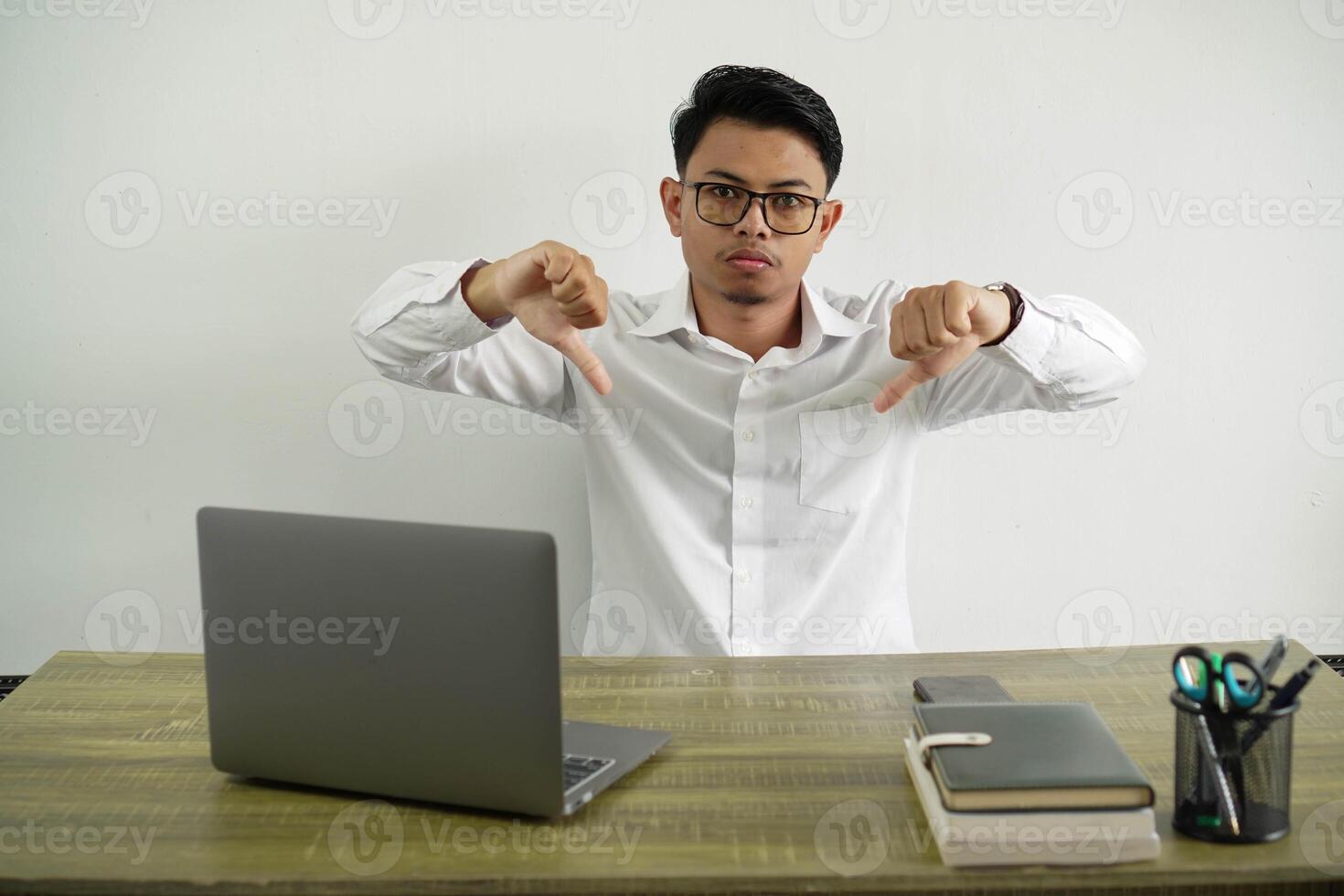 joven asiático empresario en un lugar de trabajo demostración pulgar abajo vestir blanco camisa con lentes aislado foto