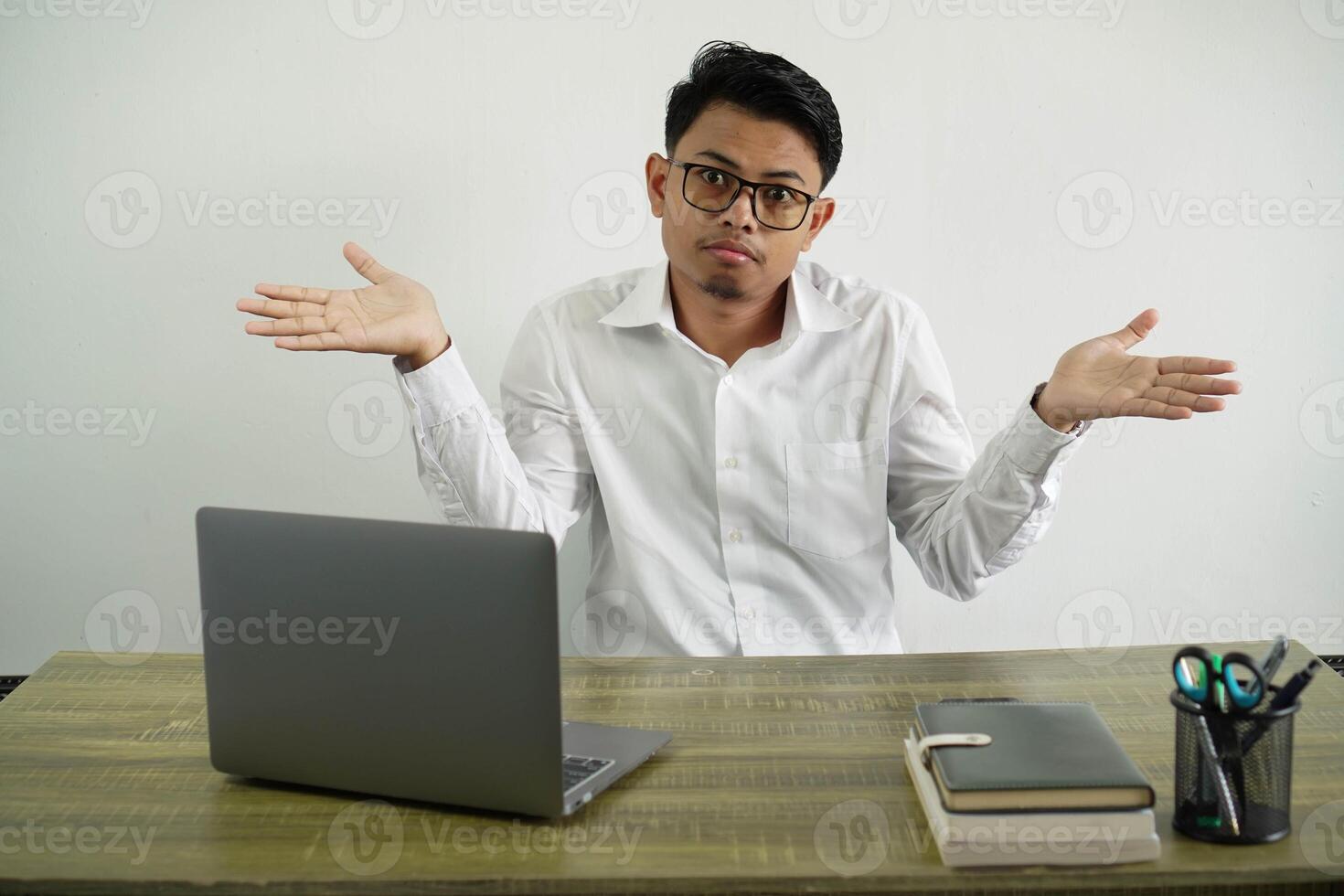 young asian businessman in a workplace having doubts while raising hands wearing white shirt with glasses isolated photo