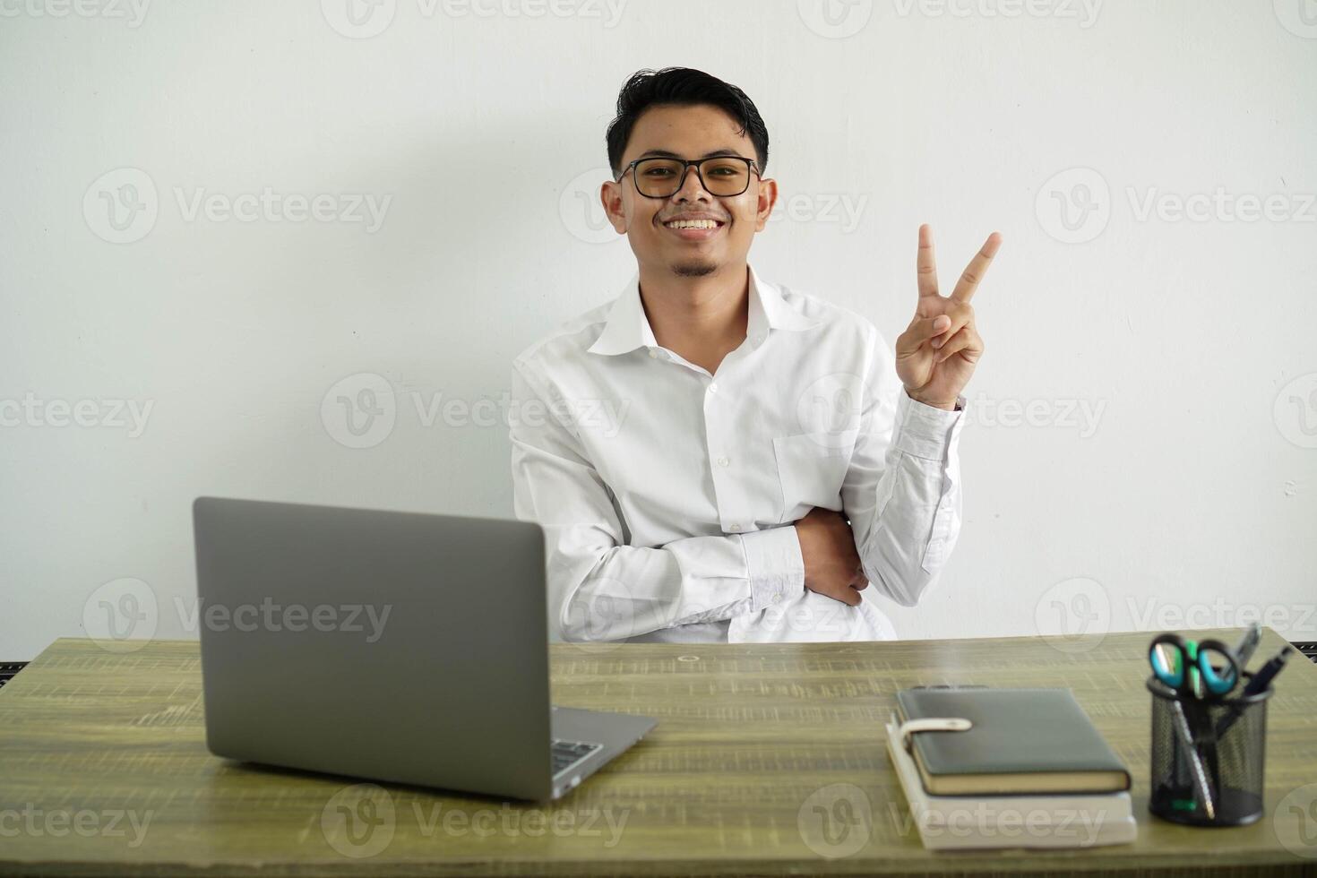 young asian businessman in a workplace smiling and showing victory sign, wearing white shirt with glasses isolated photo