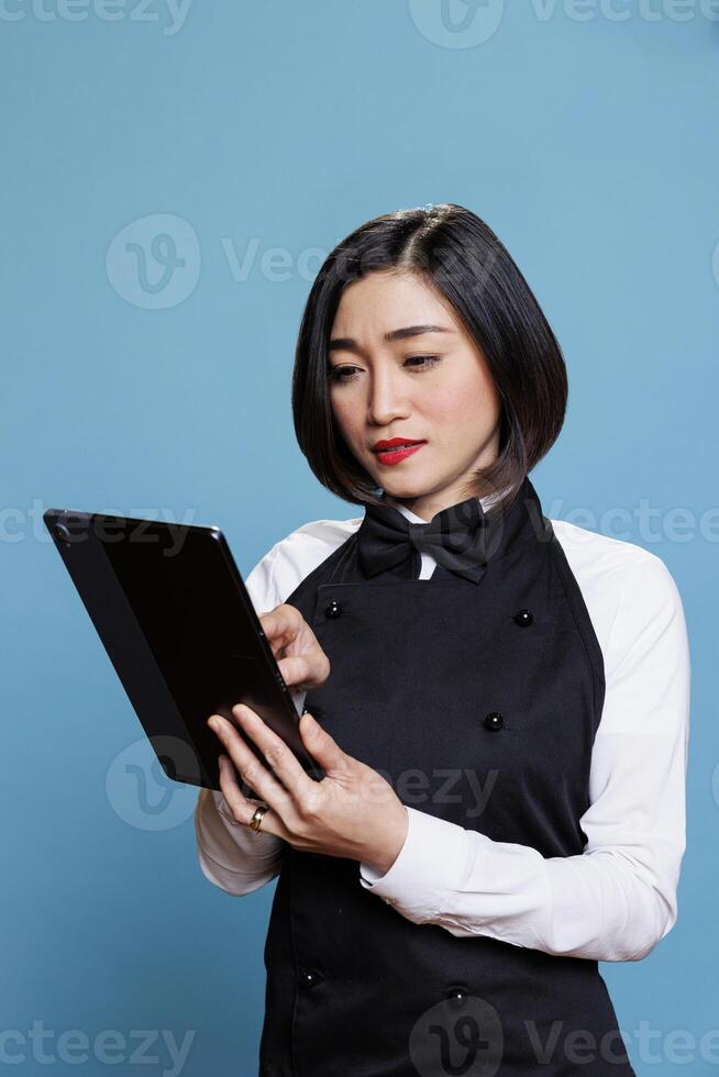 Restaurant young asian woman employee wearing uniform tapping on digital tablet. Cafeteria attractive waitress answering customer online message on portable gadget in studio photo