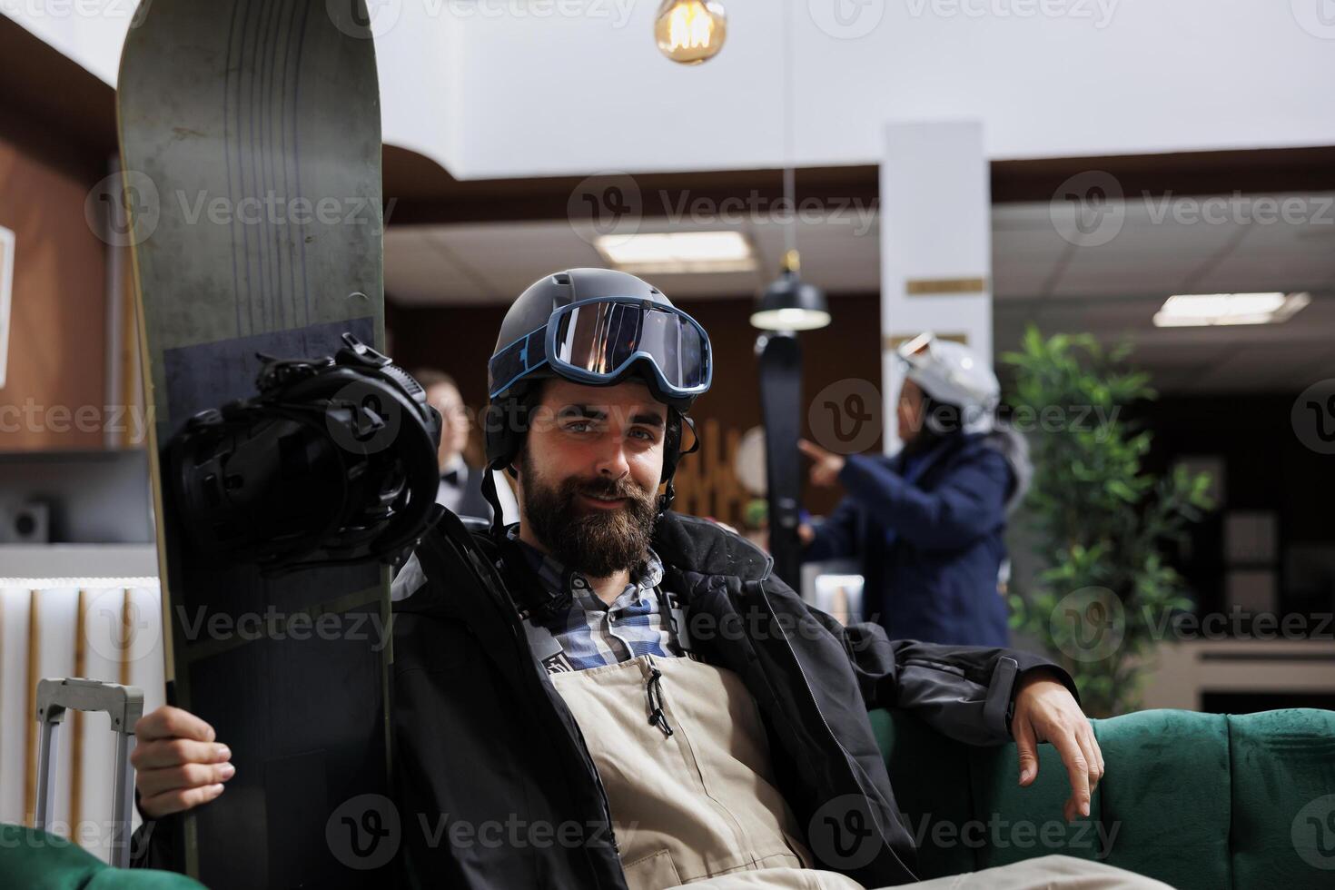 Portrait shot of caucasian male tourist wearing snow gear and holding snowboard in hotel lobby. Image showing man with winter clothing sitting in ski resort lounge with snowboarding equipment nearby. photo