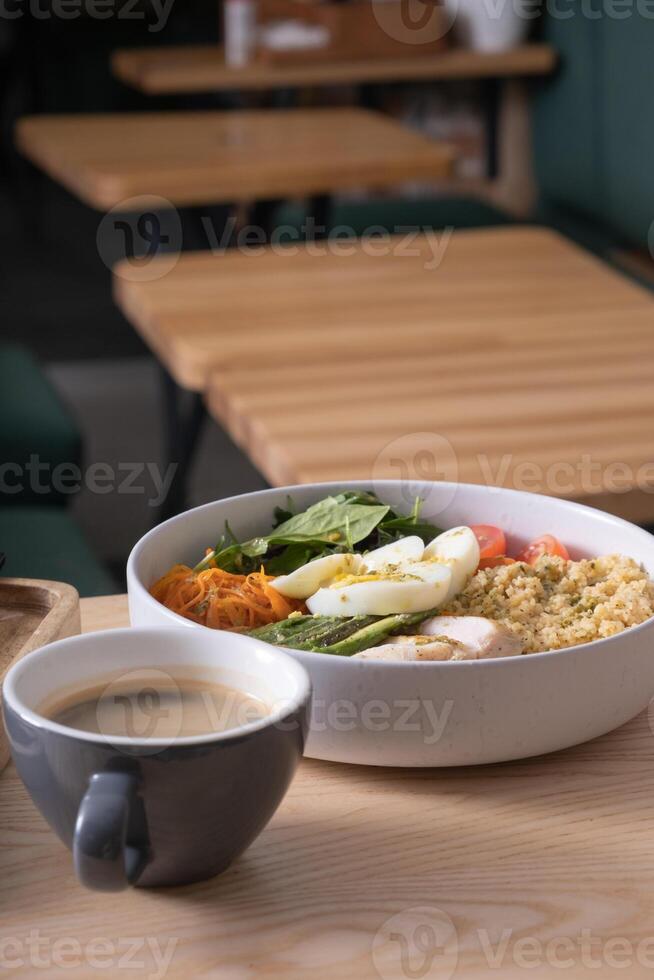 A bowl of couscous, meat, eggs and avocado and vegetables and a cup of coffee on cafe table. photo