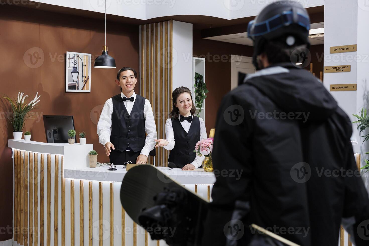 At front desk of exclusive winter resort, two receptionists warmly welcoming male guest in snow gear. Tourist wearing winter jacket and carrying snowboarding equipment arrives in hotel lobby. photo
