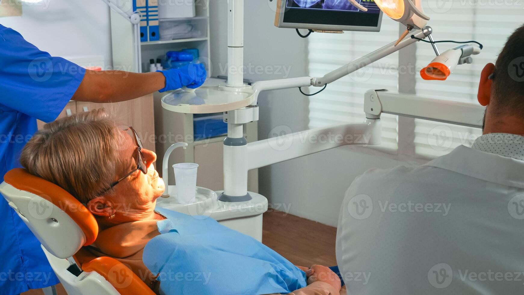 Old woman at dentist having dental treatment in implantology office. Orthodontist lighting the lamp and speaking to patient sitting on stomatological chair while nurse preparing tools for surgery. photo