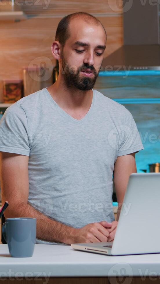 Man discussing on video call working from home sitting in kitchen looking on laptop. Busy focused employee using modern technology network wireless doing overtime for job reading writing, searching photo