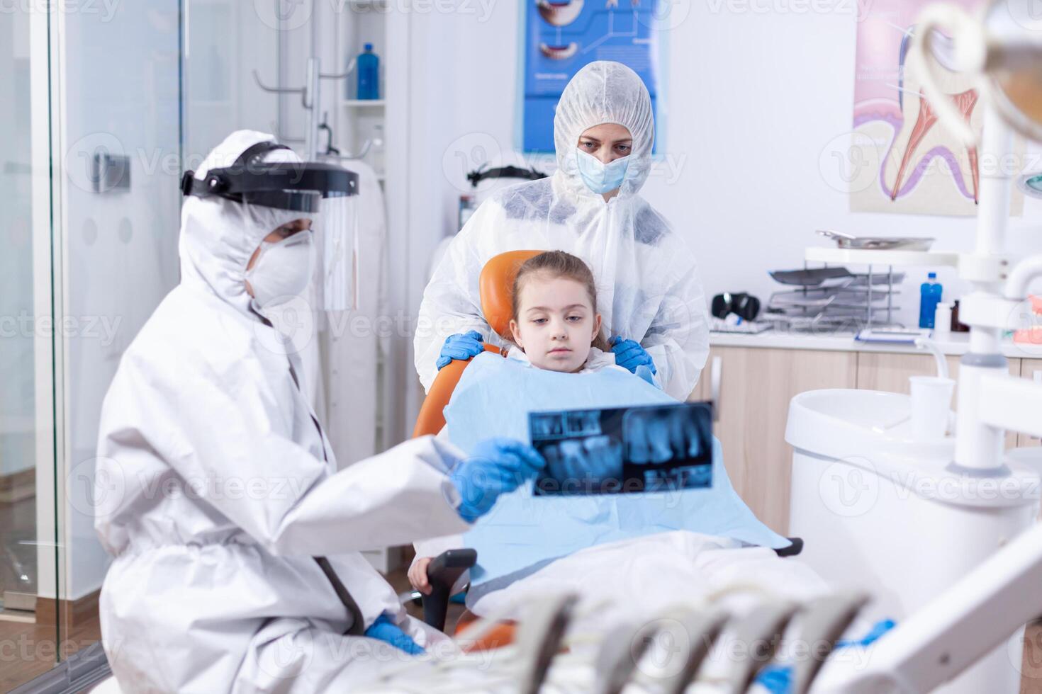 Orthodontist in ppe suit using dental radiography to explain dentistiry procedure for mouth hygine. Stomatolog in protectie suit for coroanvirus as safety precaution holding child teeth x-ray during consultation. photo