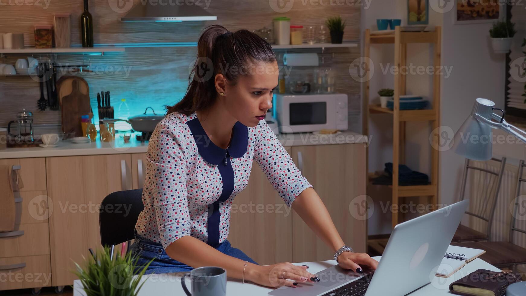 Concentrated freelance woman working overtime to finish a project from home kitchen. Busy focused employee using modern technology network wireless doing overtime for job reading typing, searching photo