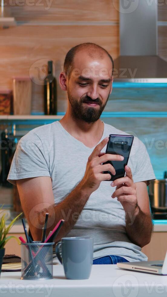 Freelancer taking break and searching on phone sitting on kitchen table working from home. Busy focused employee using modern technology network wireless doing overtime for job reading writing photo