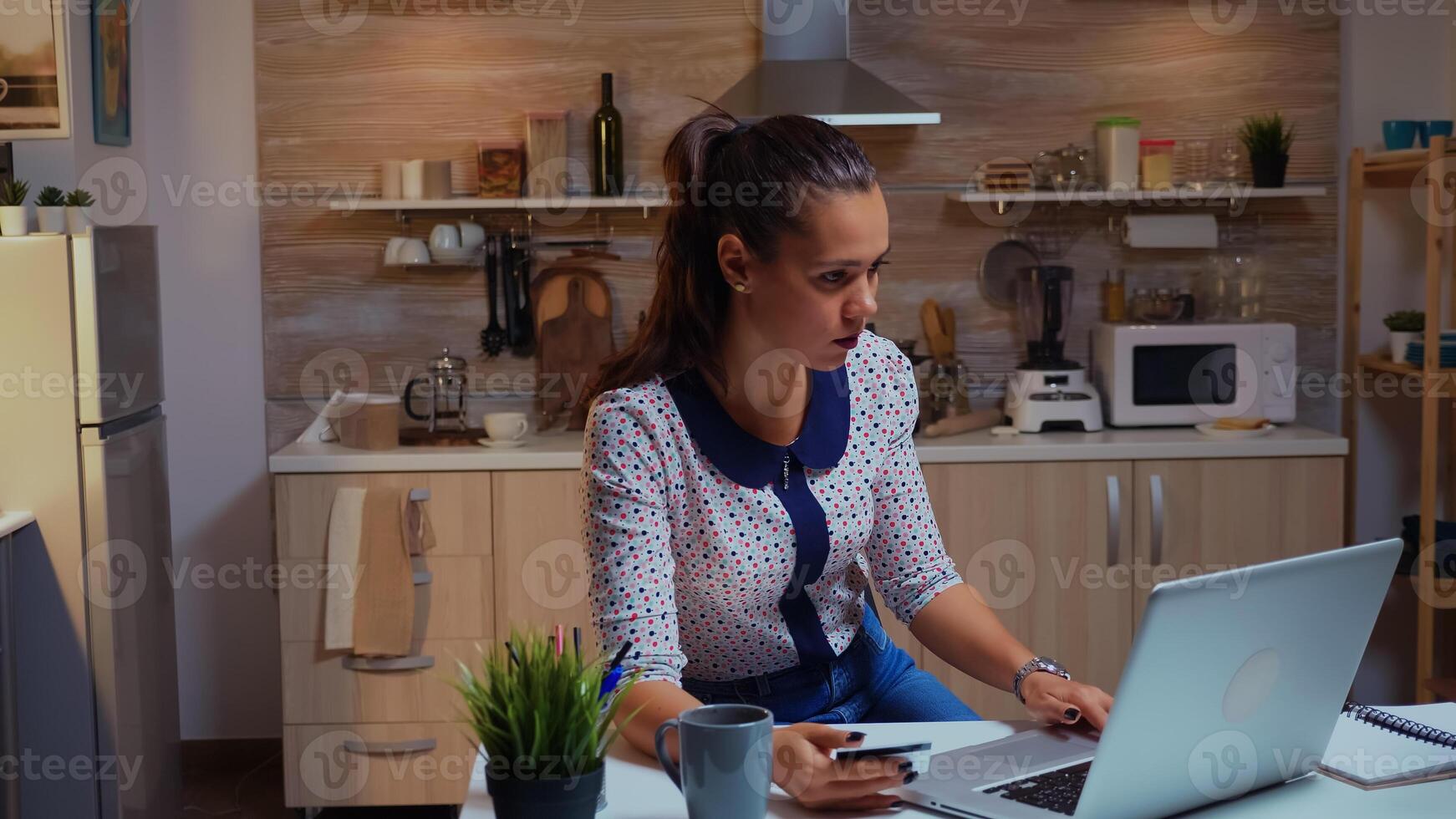 Hispanic lady holding credit card while doing online transaction using laptop in home kitchen late at night. Freelancer shopping online with electronic paymant on digital notebook conected to internet photo
