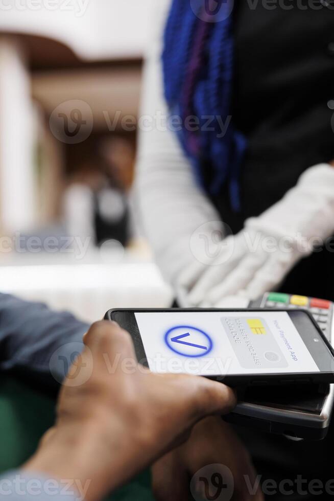 Male hotel guest making cashless payment using smartphone, phone and card reader machine, close up. Tourist paying for services with phone. Waiter accept contactless NFC payment from customer photo