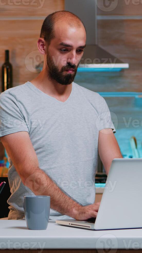 Tired sleepy man yawning, working at home desk in the kitchen. Busy overwork focused employee using modern technology network wireless doing overtime for job reading writing, searching photo