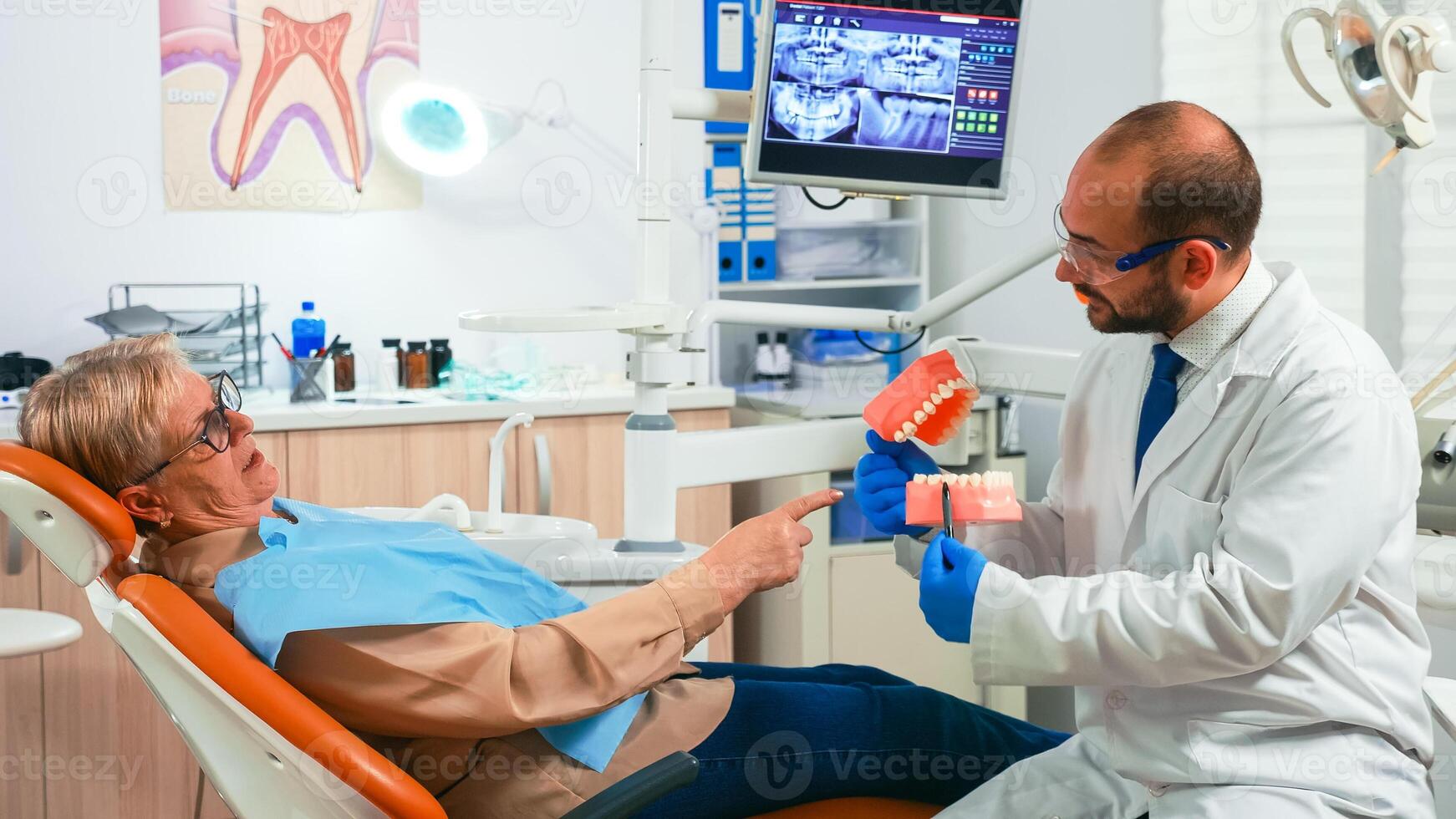 Young stomatologist explaining to senior patient procedure using model of dental teeth. Doctor holding sample of human jaw telling informations for keeping healthy teeth, digital x-ray in background photo