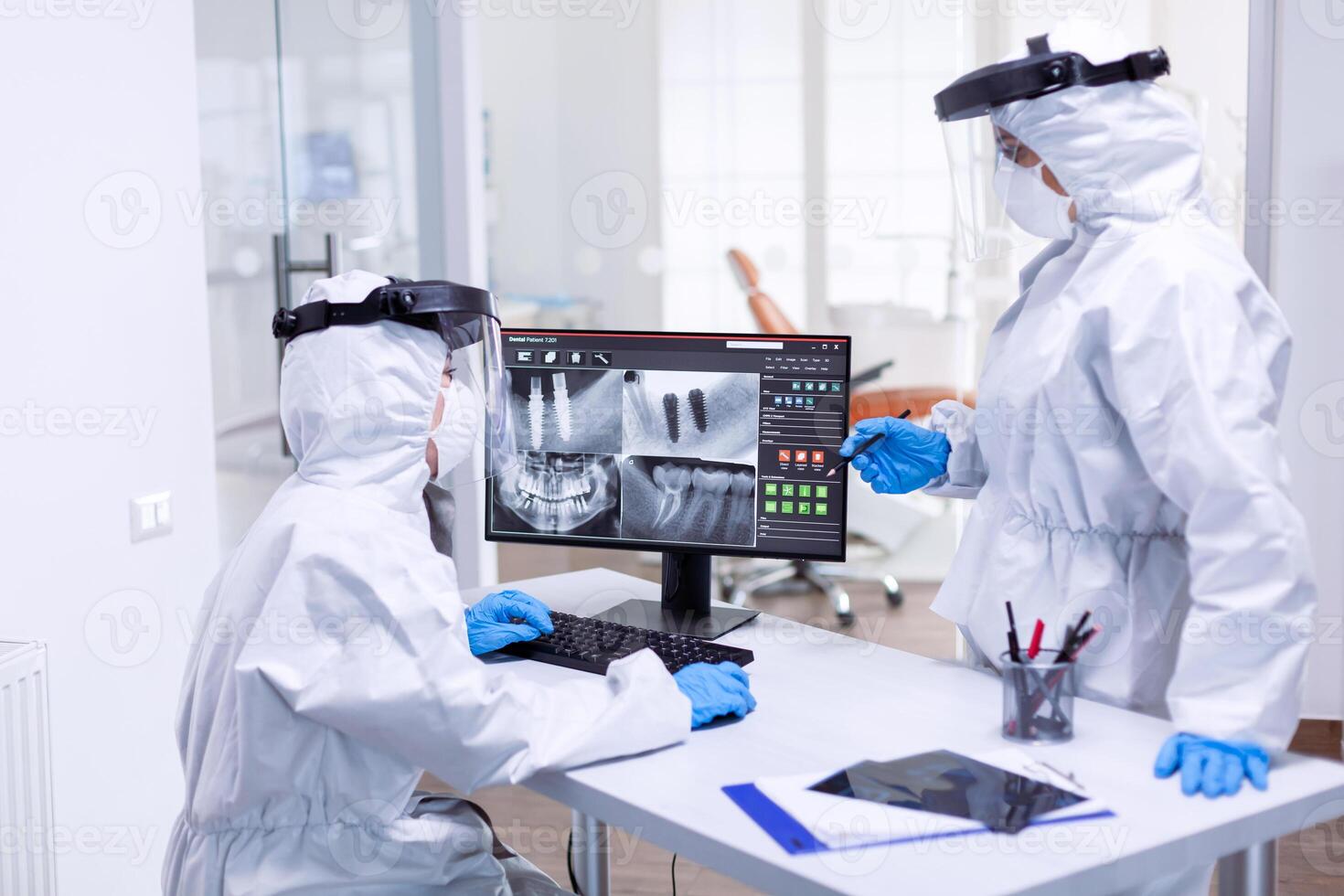 Dentist and nurse discussing patient teeth issues dressed in ppe suit. Medical specialist wearing protective gear against coronavirus during global outbreak looking at radiography in dental office. photo