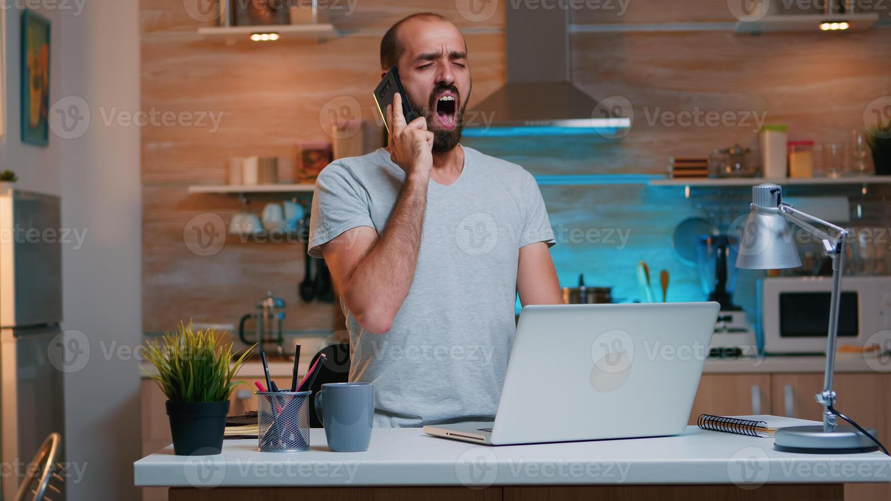 Tired freelancer talking on phone yawning sitting at kitchen desk in on laptop late at night. Busy employee using modern technology network wireless doing overtime for job reading writing, searching photo