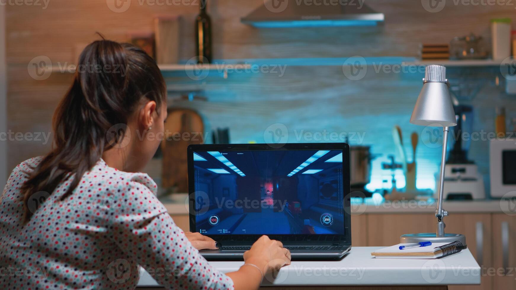 Woman gamer sitting at home in kitchen testing new game on professional laptop. Tired player gaming online video games on her personal computer with modern technology network wireless photo