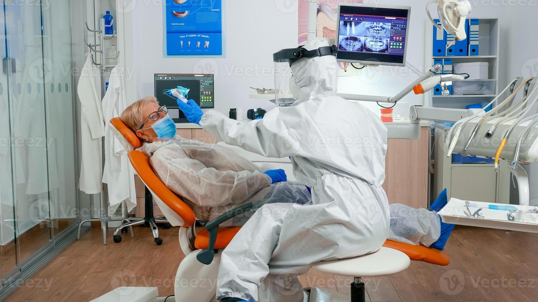 Dentist assistant in potective suit measuring patient temperature before dental examination during coronavirus pandemic. Nurse in coverall and mask interrogating woman and taking notes on clipboard photo