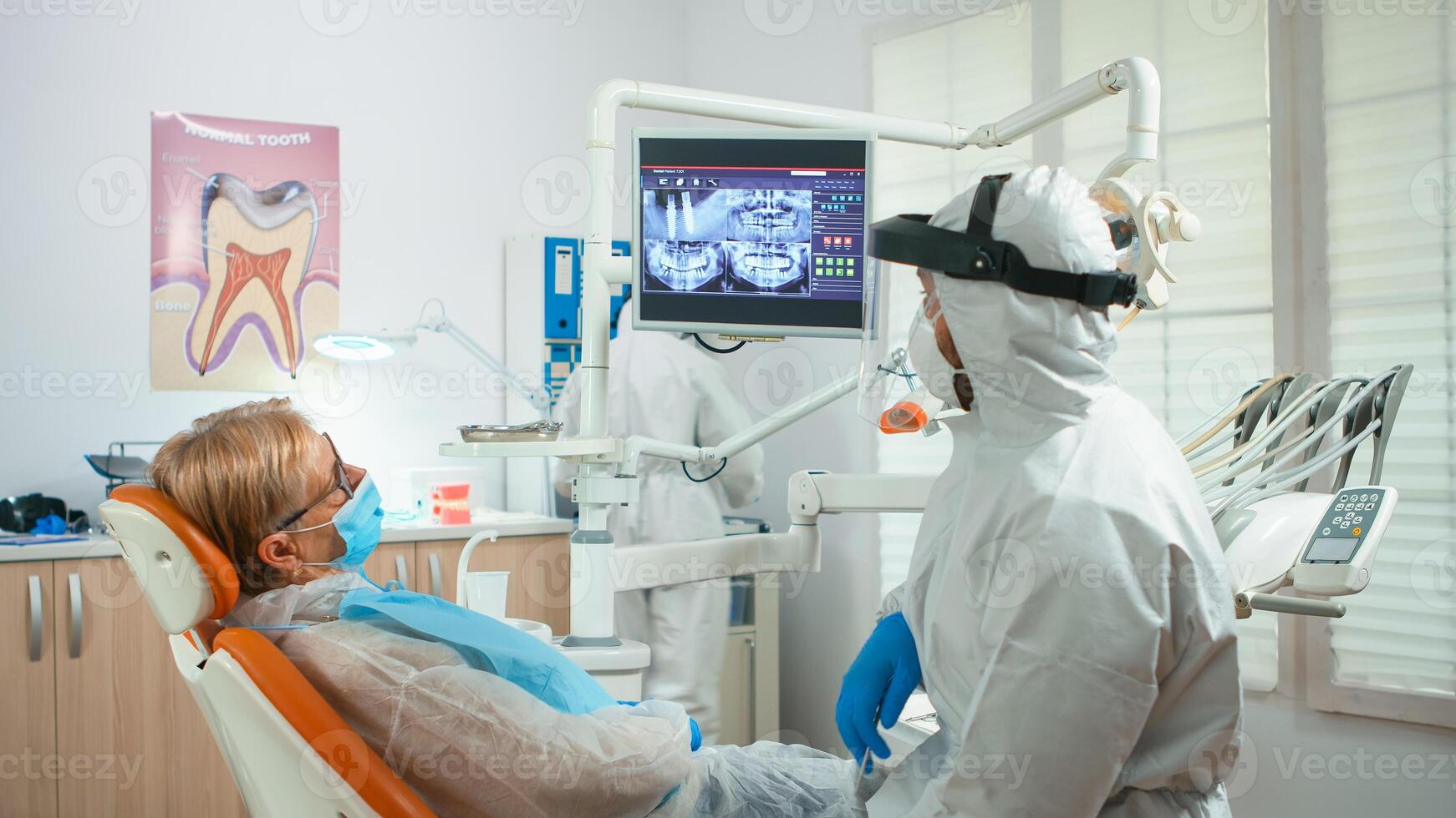 Orthodontist in special equipment pointing on digital x-ray explaining dental treatment during global pandemic. Medical team talking with woman wearing face shield, protection suit, mask and gloves photo