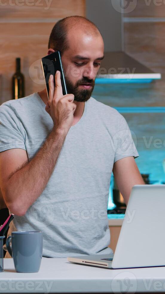 Remote employee speaking at phone while working at laptop typing late at night. Busy focused freelancer using modern technology network wireless doing overtime for job reading writing, searching photo