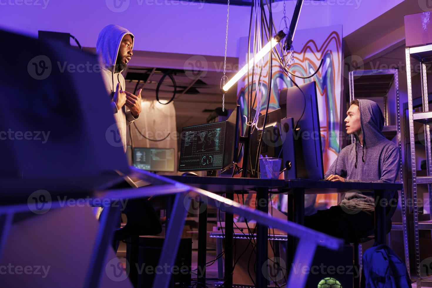 Two diverse hackers working together to hack computer system in abandoned warehouse. Asian and african american internet criminals discussing malicious software code in dark workspace photo