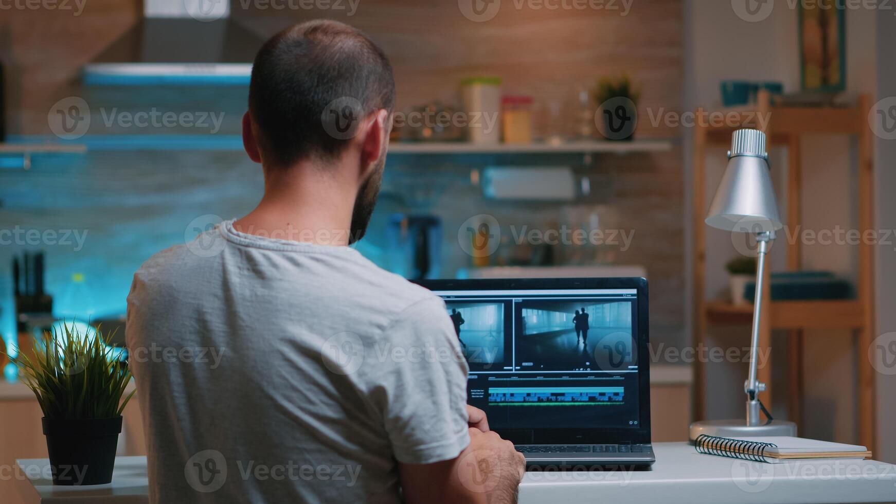 Colorist working remotely overtime from home using editing software sitting in front of laptop late at night. Videographer processing audio film montage on professional in modern kitchen in midnight photo