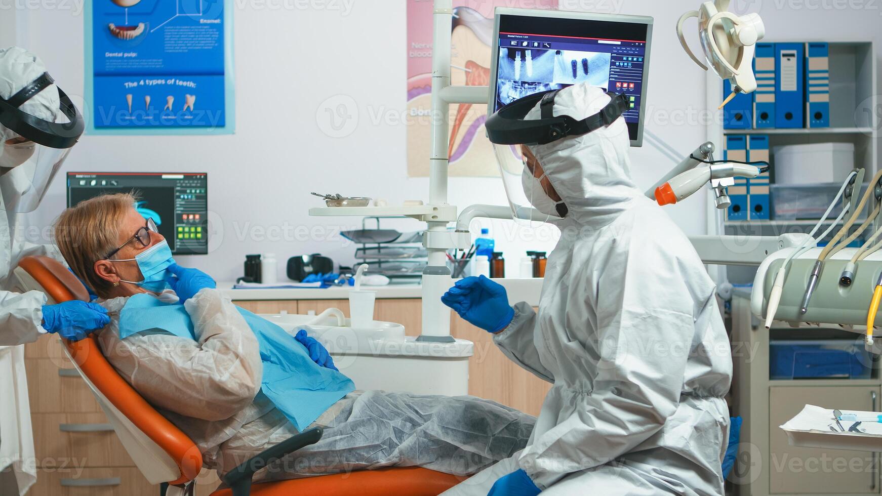 Dentist doctor with protection equipment talking with old patient before consultation during covid-19 epidemic. Concept of new normal dentist visit in coronavirus outbreak wearing protective suit and face shield photo